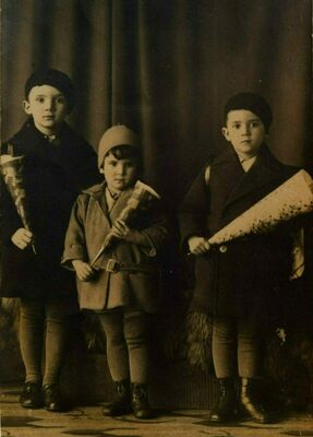 Trois écoliers vêtus de bérets et de manteaux posent devant de sombres rideaux. Chacun tient un cornet de papier.