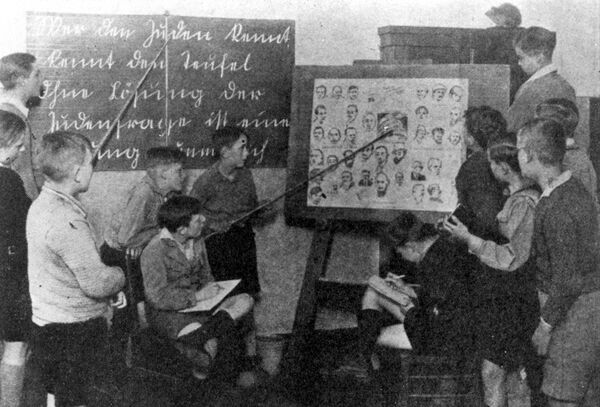 Children crowding around an easel, one of them holding a pointer up to sketches of faces and heads, in the background, a boy holding a pointer up to a blackboard with German writing on it.
