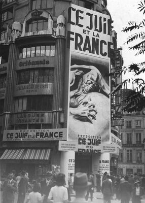People milling outside a building featuring a large poster on its façade with French writing and a caricature of a man's face bent toward planet Earth and one of his hands grasping at its surface.