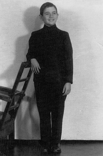 A boy in black formal clothing standing against a light background, gazing off to the side and smiling, his right arm resting on the back of a chair.