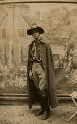 A boy with a serious expression, wearing a long cape and a hat with a wide brim, standing in front of a wall featuring a mural of a house and flower garden.