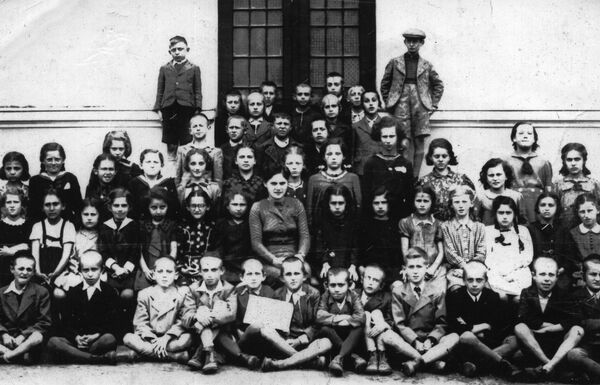 Filles et garçons sont installés devant un bâtiment pour la photo de classe. Leur enseignante est assise au centre.