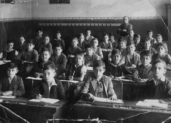 Classroom full of young boys sitting at desks with books open in front of them, looking at the camera, their teacher standing at the back of the classroom with his arms folded.
