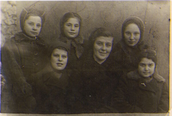 Cinq fillettes vêtus de chapeaux et de manteaux posent avec une femme. Elles affichent de timides sourires.