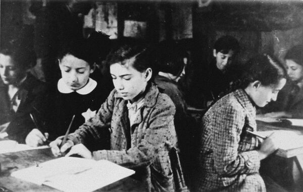 Children sitting at tables with books open, writing, some wearing coats, a Star of David visible on one child's clothing.