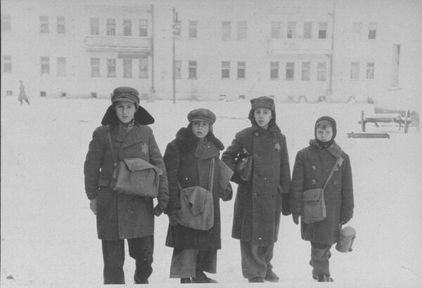 Children standing outside in snow, wearing hats and coats with Stars of David on them, carrying bags worn across their bodies or held under their arms, a large building in the background.