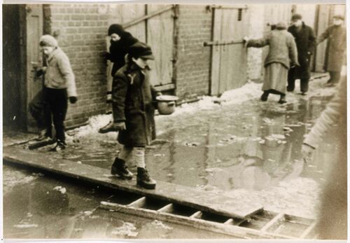 <p>A child holding a soup bowl crosses a flooded street on a plank of wood in the Lodz ghetto. Lodz, Poland, circa 1940–1944. </p>
<p><em>United States Holocaust Memorial Museum, courtesy of Arie Ben Menachem.</em></p>