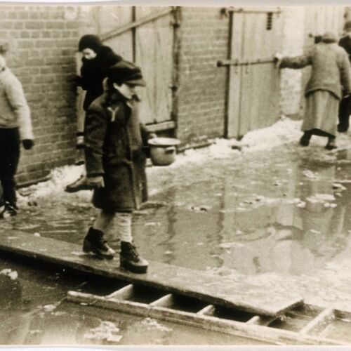 <p>A child holding a soup bowl crosses a flooded street on a plank of wood in the Lodz ghetto. Lodz, Poland, circa 1940–1944. </p>
<p><em>United States Holocaust Memorial Museum, courtesy of Arie Ben Menachem.</em></p>