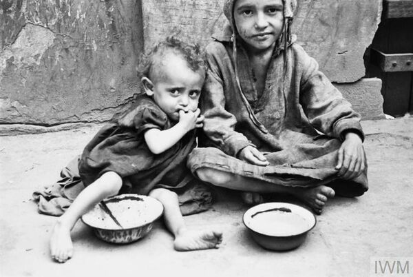 Deux enfants maigres en haillons sont assis près de leurs bols posés à même le sol. Le plus jeune enfant appuie sa tête sur l’épaule du plus grand.