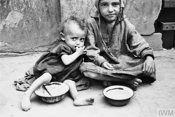 Two very thin young children sitting on the ground, barefoot, wearing ragged clothing, the younger child leaning against the older one, bowls on the ground in front of them.