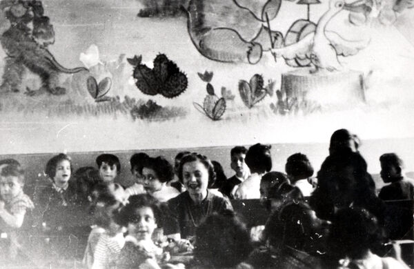 A smiling woman and children sitting at long tables with a cartoon mural partially visible on the far wall.