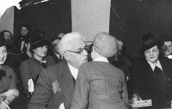A young child with their back to the camera leaning toward a man with white hair, with a Star of David visible on his jacket, several women looking on and smiling.
