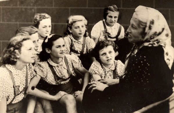 A group of girls with intent looks on their faces, sitting and leaning toward a woman with a kerchief who is sitting in front of them, Stars of David visible on some of their clothing.