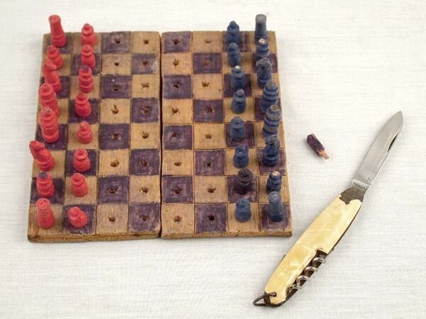 A chess set made of a wooden board with holes in the squares and red and blue pieces, a penknife open next to it.