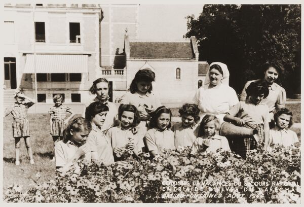 A group of girls stand behind a flowering hedge with a woman in a white habit, two girls looking on in the background and a building behind them.