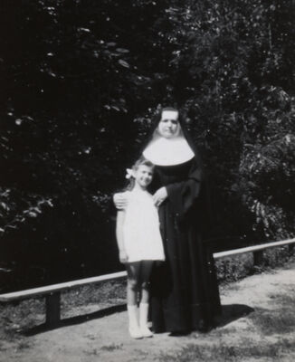 A nun in a habit standing protectively next to a smiling young girl, hands on her shoulders.