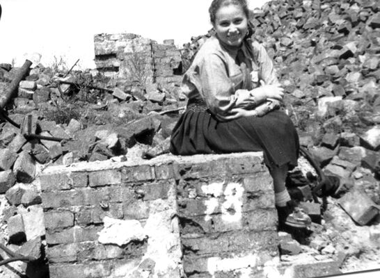 Smiling girl sitting on remnant of brick wall with piles of brick and rubble all around her.