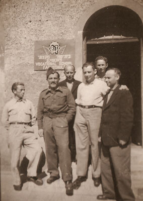 People posing at the entrance to a building and a sign with Yiddish, English and German writing on it.