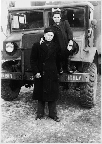 A large jeep with the words UNRRA and Italy written on the front bumper, one child standing in front of it, another standing on the bumper with her arm around the other child.