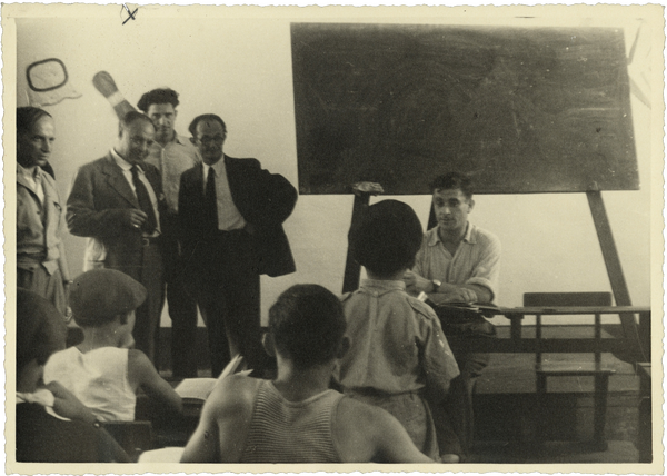 A group of men looking on as a teacher sits in front of a classroom with several students, a blackboard behind him.