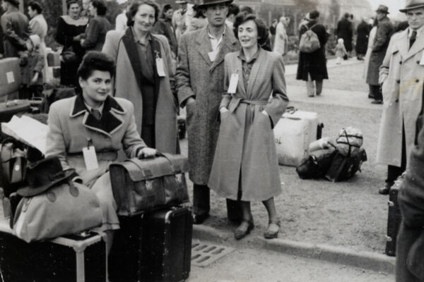 A woman sitting with a pile of luggage, looking at the camera, several people standing behind her with luggage and people milling in the background.