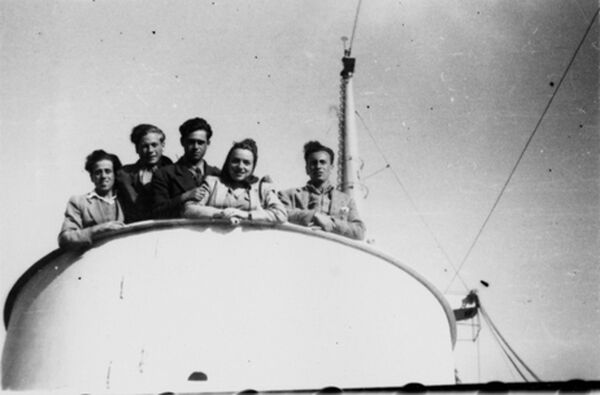 Several young people leaning over the edge of a rounded barrier, looking down at the camera, the mast of a ship behind them.