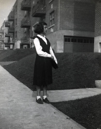 A girl standing on a sidewalk outside a row of low-rise apartment buildings.