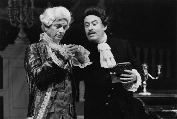 Two men in period costumes and wigs examining something they are holding.