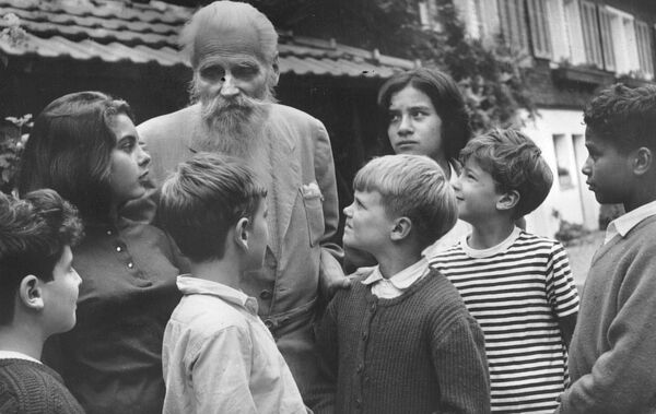 Man with white hair and beard with group of children standing around him and looking up at him.