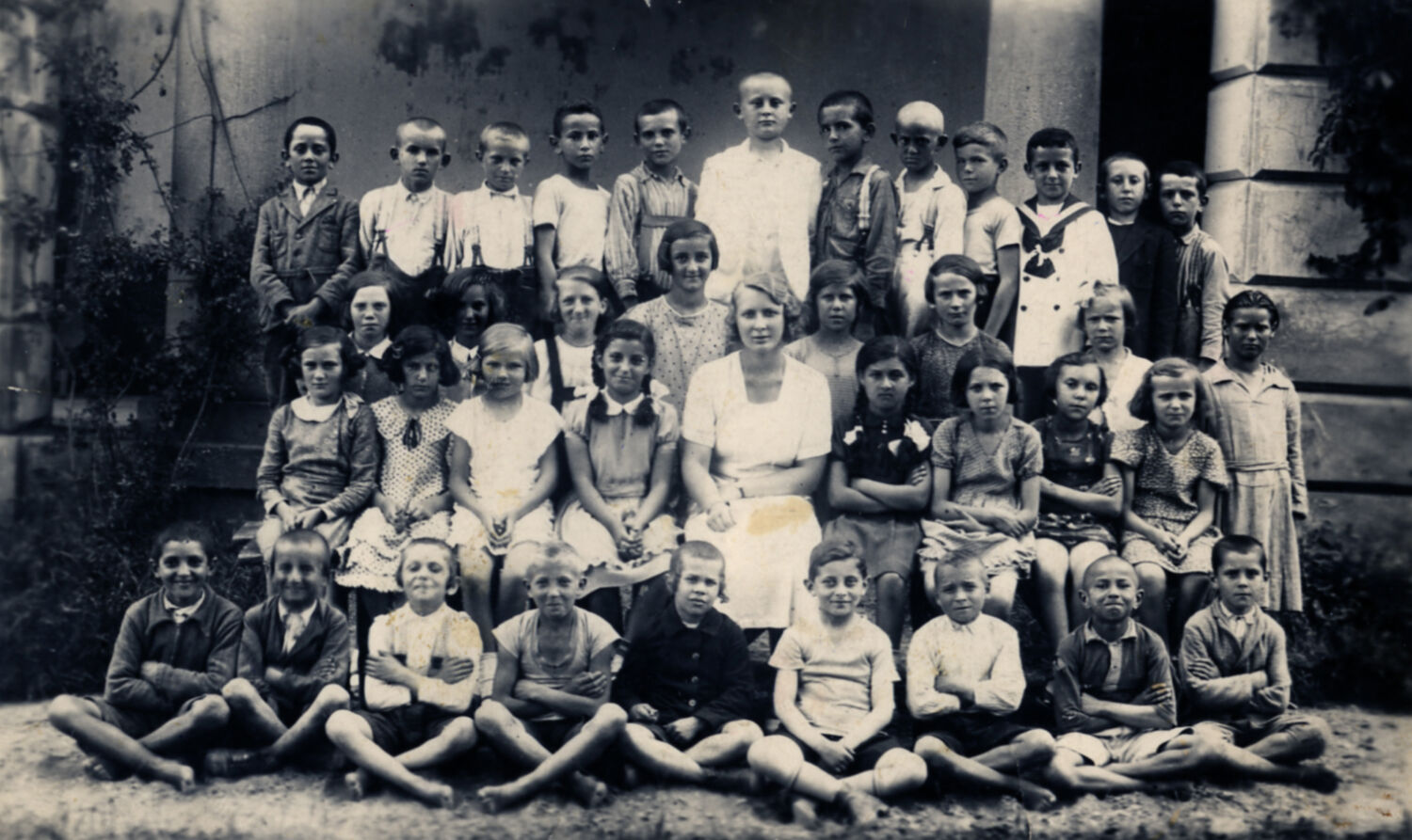 A large group of children posing for a class photo with their teacher in front of a building.