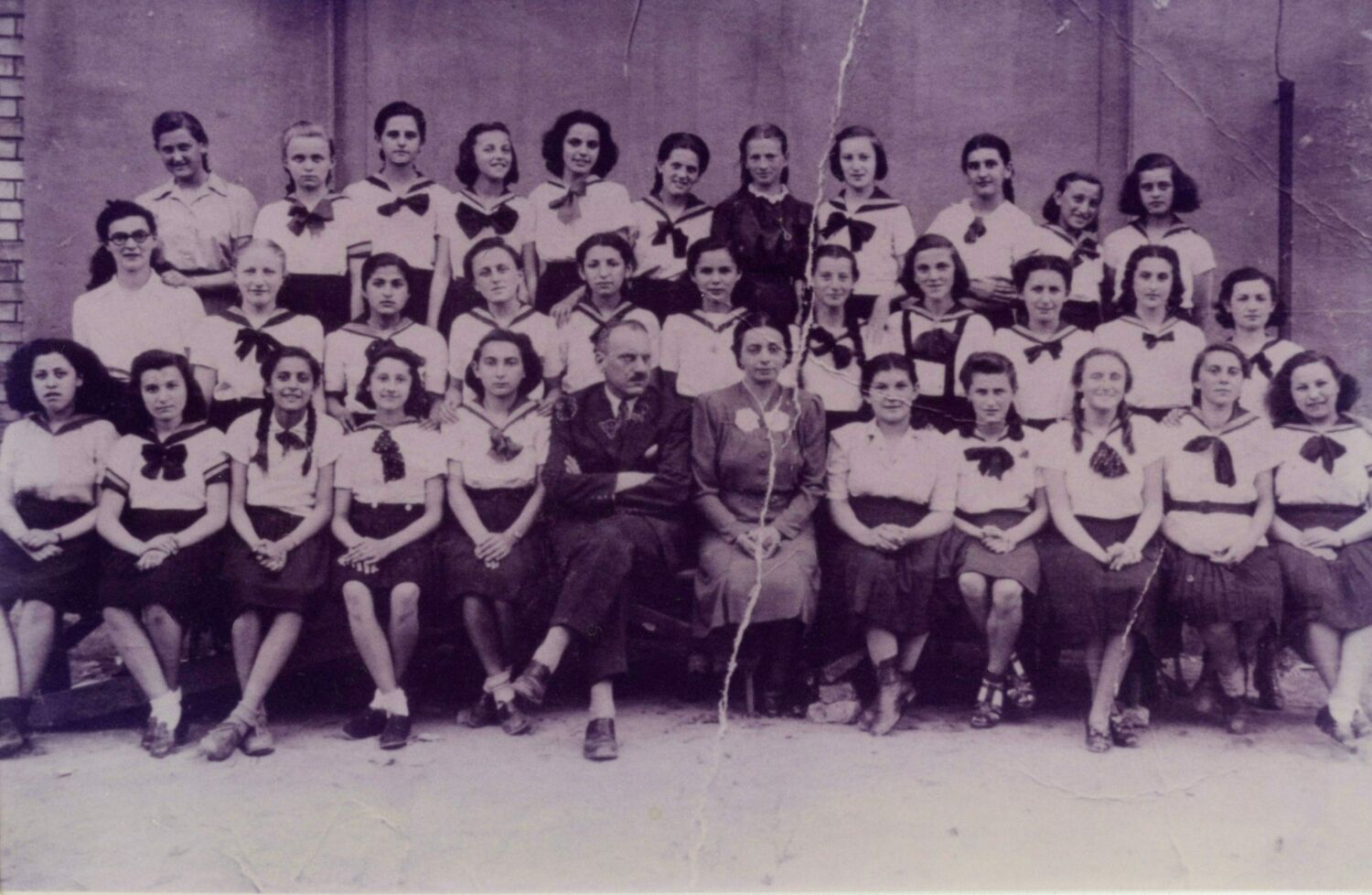 A class photo of girls wearing a uniform of a white top and dark skirt, two teachers sitting in the front row.