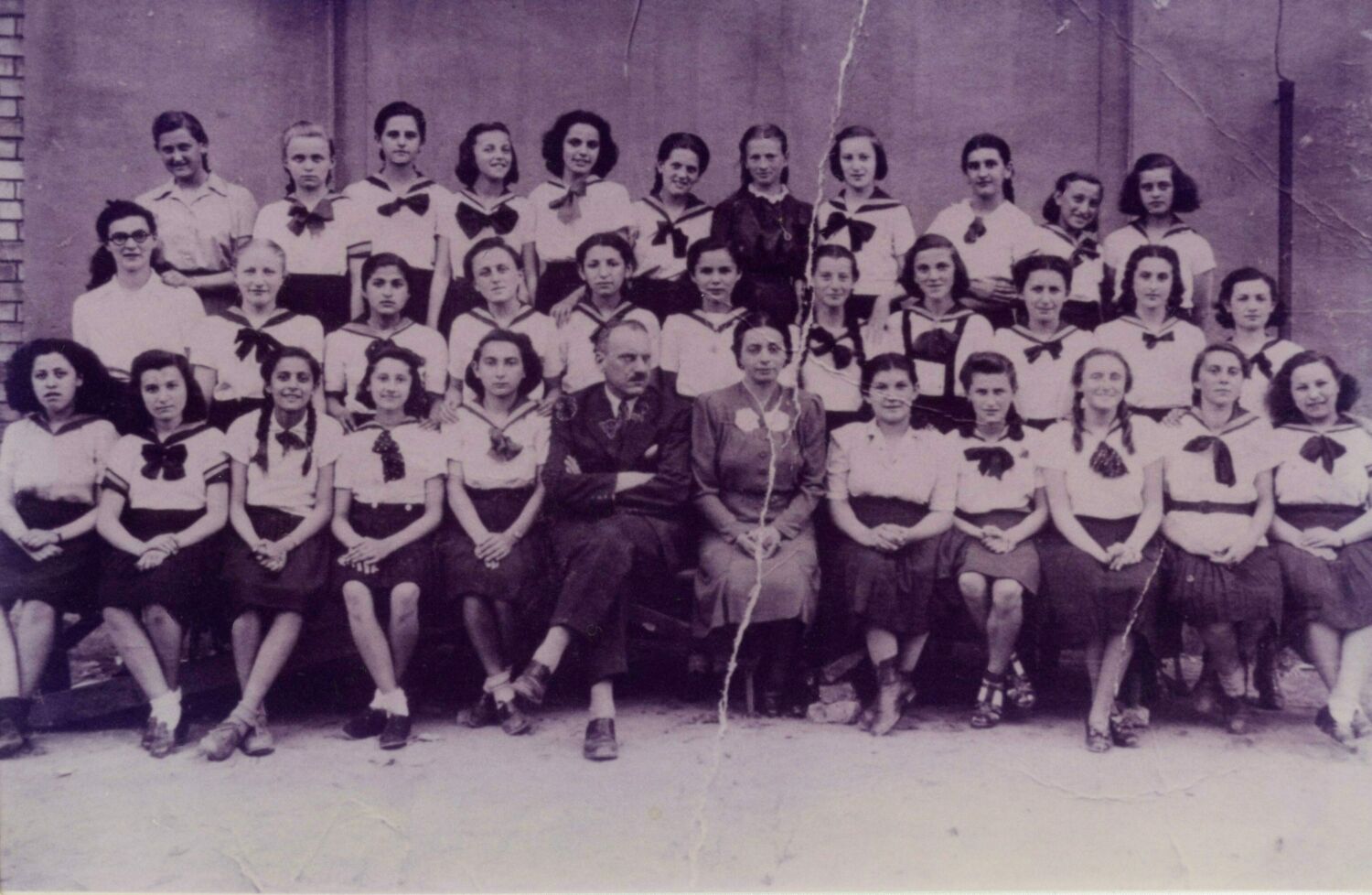 Une photo de classe d’adolescentes en chemises blanches et en jupes noires. À l’avant, deux enseignants ont un air sérieux.