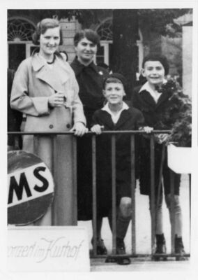 Derrière une clôture, deux femmes et deux garçons sourient. On décèle des panneaux en allemand fixés aux barreaux.