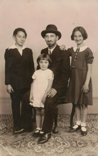 Un homme vêtu d’un chapeau est assis. Une fillette se tient debout devant lui et deux autres enfants, à ses côtés.