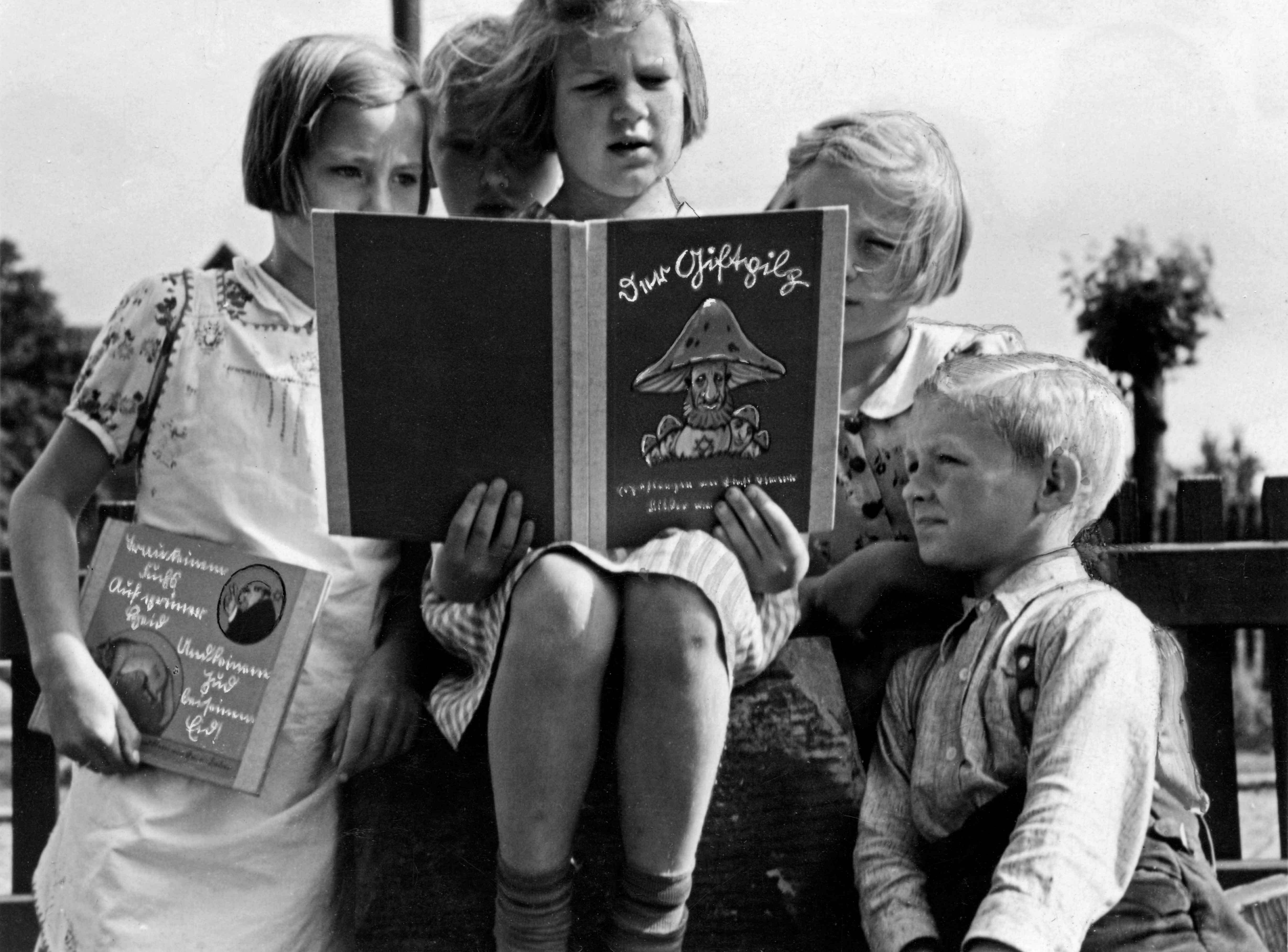 A group of children looking at a book with a cover featuring German lettering and a caricature of a face with a mushroom hat.