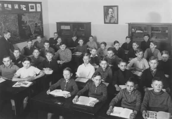 Dans une classe bondée, des garçons sont assis à leur pupitre devant leurs livres ouverts. Un portrait de Hitler est suspendu au mur du fond.