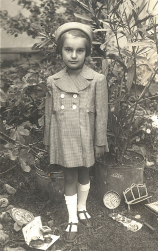 Une jeune fille vêtue d’un chapeau et d’un manteau pose dans un jardin. Des jouets sont dispersés à ses pieds.