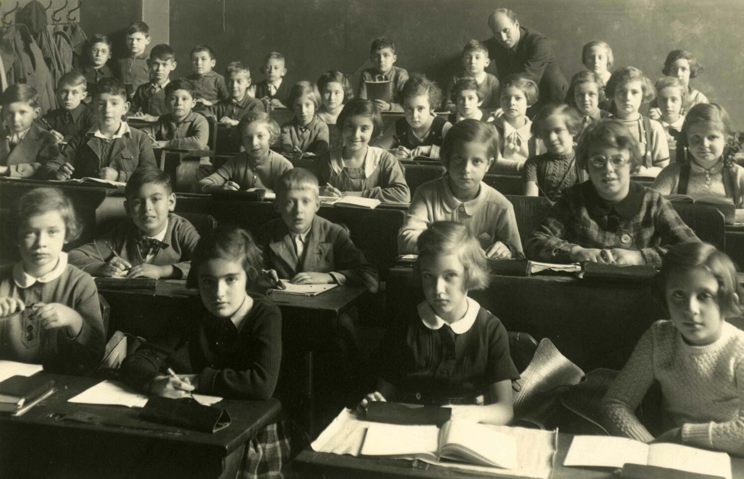Young children sitting at desks with books open, looking at the camera, a teacher leaning over one child's desk at the back of the classroom.
