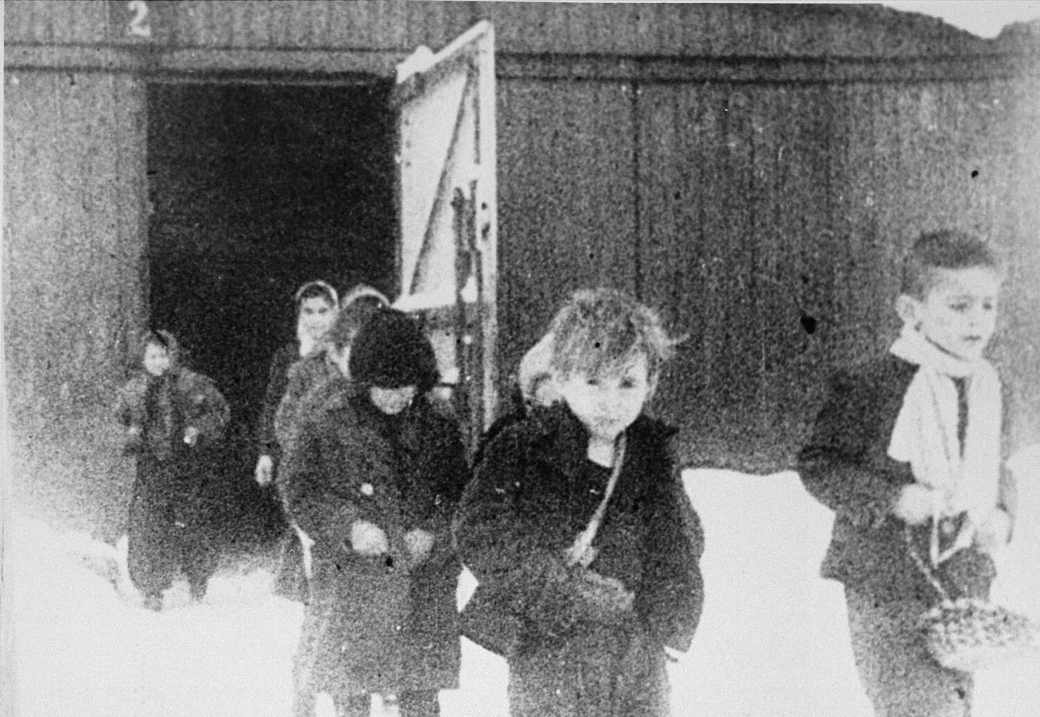 Young children in jackets and scarves walking out of a large door in a wooden building, snow on the ground.
