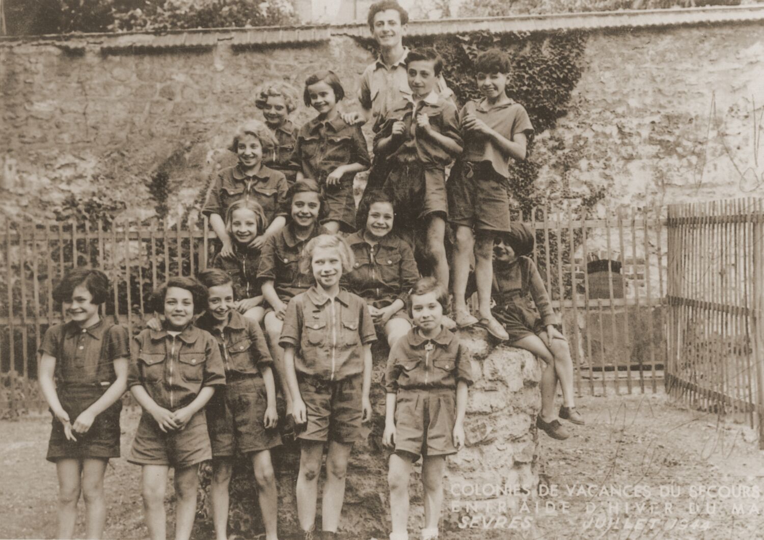 A group of children looking happy and mischievous, standing against and on top of a rocky platform outside with an adult.
