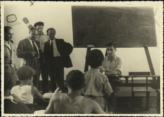 Des hommes observent une classe en action : un enseignant est assis devant un tableau noir et des élèves sont installés à leurs pupitres.