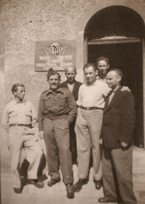 Six hommes posent devant un immeuble sur lequel est fixé un écriteau avec les lettres « O R T » et du texte en yiddish, en anglais et en allemand.