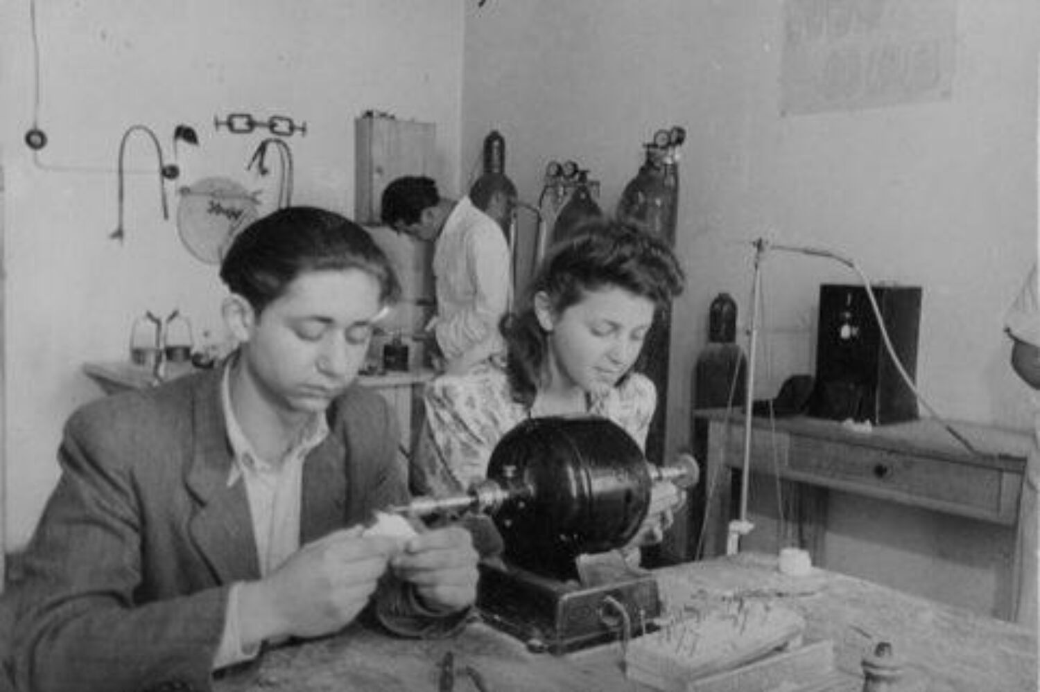 Boy and girl sitting next to each other at a table with a small machine on it, focusing intently on items in their hands, a person in a lab coat working in the background.