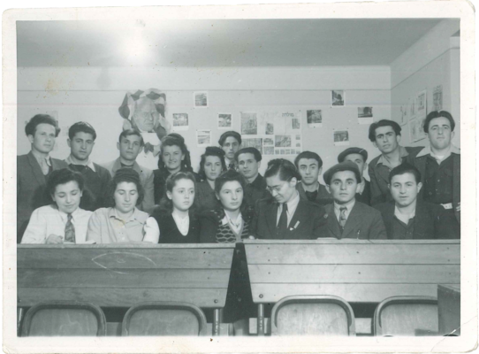 A group of teenagers sitting and standing close to each other behind desks in a classroom.