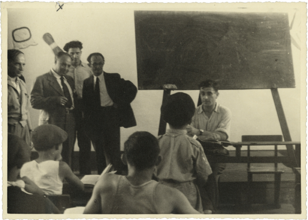 A group of men looking on as a teacher sits in front of a classroom with several students, a blackboard behind him.