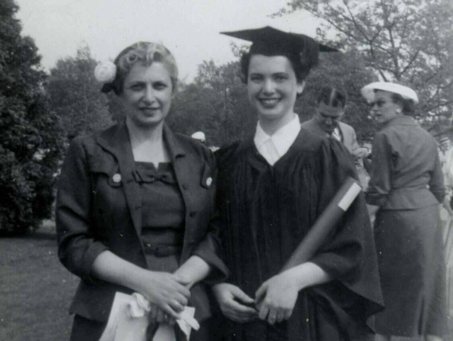 Two women standing close to each other outdoors and smiling, one of them wearing a cap and gown.