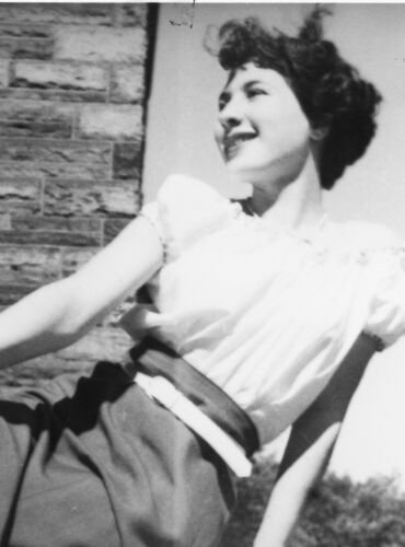 A black-and-white photograph of a young woman posing outside of a stone building. Her dark short hair is styled at ear-length. She is looking off to the left with a smile. She is wearing a white short-sleeved blouse, with a long grey skirt and a belt.