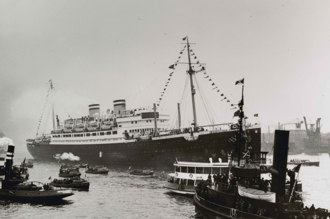 Black-and-white photograph of a large ship in the water with flags on two masts and covering lines from mast to deck. Smoke is billowing from the ship’s two smokestacks.